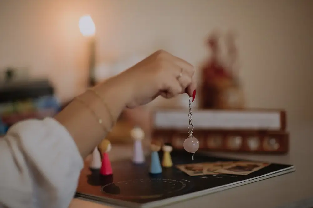 Mão de Debora segurando um pêndulo de quartzo rosa sobre um gráfico radiônico, com cartas de tarô ao lado